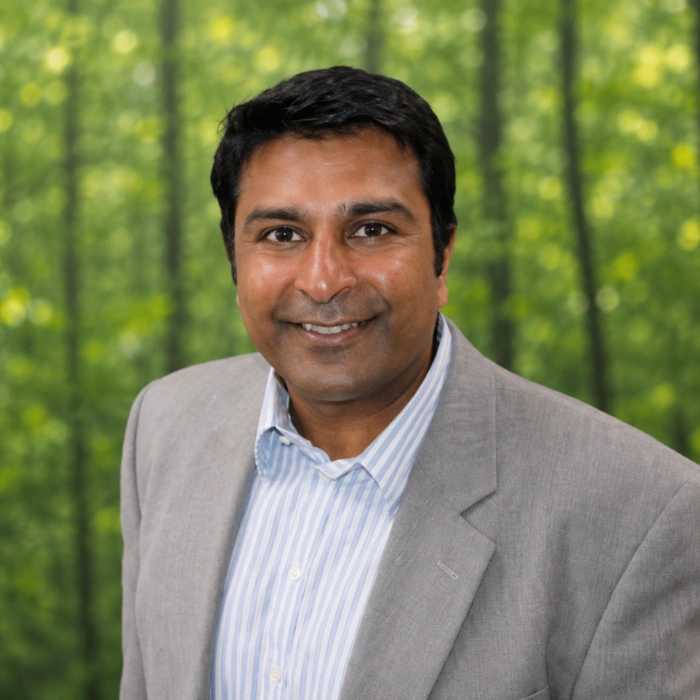 Nimish Shah Headshot. He is smiling in a gray suite with a white shirt posed in front of trees.